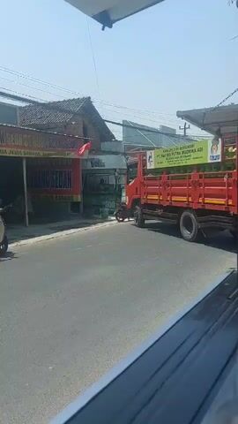 A red truck is parked on the side of a road. The truck is loaded with green objects, possibly watermelons. In front of the truck, a sign displays text in Indonesian. To the left of the truck, there is a building with a red and white flag. A motorcycle with two people is seen riding on the road. The sky is clear and blue, indicating a sunny day.
