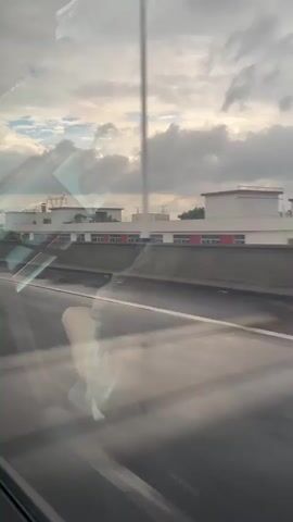 The video shows a view from inside a vehicle, looking out a window at a highway and buildings in the distance. The sky is cloudy with hints of sunlight breaking through. Reflections are visible on the window, obscuring some of the details of the scene. In the foreground, a person's leg and arm are visible, suggesting they are sitting in the passenger seat. The highway appears to be empty of other vehicles. The buildings in the distance are low-rise structures with red accents. Electrical towers are also visible in the background. The video is shaky, indicating movement of the vehicle.