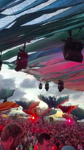 The video shows a large outdoor rave. A crowd of people stand facing a stage, underneath a canopy of blue, green, pink, and red fabric hung from the ceiling and resembling giant tentacles. There are black lights and speakers attached to the canopy. The sky behind the stage is partially visible and filled with clouds. The event appears to be taking place at dusk.