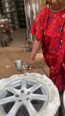 A woman is shown spray-painting a car wheel silver. She is wearing a red and blue floral print dress. The wheel is partially covered with masking tape. A person is partially visible behind her with their hand on her shoulder. The background shows a collection of car wheels and a building with signs. The video focuses on the woman's activity with the spray gun and the wheel.