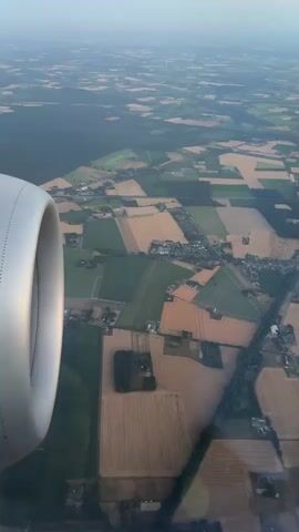 This is a vertical video taken from an airplane window. On the left side of the frame is the engine nacelle of the airplane. Below the airplane, a landscape of agricultural fields can be seen. The fields are in various shades of green and tan, suggesting different crops or stages of cultivation. Some patches of trees and a small village or settlement are also visible. The horizon is visible in the distance, with a hazy sky. The video shows the perspective of looking down and slightly forward from the aircraft.