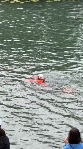 A person wearing a white cap and an orange life vest is swimming in a body of water. They appear to be pulling another person, also wearing an orange life vest, in the water. The water is green and rippled, with lily pads visible near the top of the frame. Two people are visible in the foreground on the shoreline, one wearing a white cap and the other holding a camera.