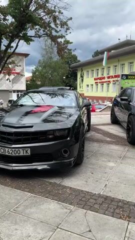 The video shows two black cars parked on a paved area in front of a building with a light yellow facade. The building has windows and a large sign with Bulgarian text. The car in the foreground is a Chevrolet Camaro, and the car in the background appears to be a Dodge Charger. The Camaro is black with red accents on the hood and a red Chevrolet logo. The Charger is also black with red stripes on the hood. Both cars have custom rims. The sky is cloudy. There are trees and a flag visible in the background.