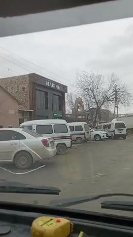 The view from inside a vehicle looking through the windshield shows a wet, potholed street lined with parked cars, vans, and a truck. A building with the name "MADINA" is visible on the left. The sky is overcast. A yellow tape measure sits on the dashboard in front of the windshield. The vehicle appears to be moving slowly forward.