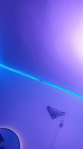 The video displays an interior space featuring modern LED lighting. A strip of blue LED lights runs horizontally along a light-colored wall. Above this, a contemporary chandelier hangs from the ceiling, consisting of three illuminated concentric rings of varying sizes, with the largest at the top and smallest at the bottom. Below the blue LED strip, a decorative, fan-shaped metallic ornament is mounted on the wall, with a white electrical fixture, possibly a light switch or outlet, positioned underneath it. A fragment of a dark, circular object, potentially a mirror or another decorative element, is visible in the bottom left corner. The room is predominantly illuminated by the blue and white LED lights, casting a purple-blue ambient glow. The camera pans slowly upwards, revealing the chandelier in full.