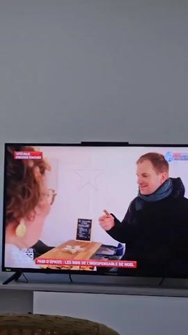 A television displays a scene from a show with the title "UN JOUR UN DOC de Noel" in the lower-left corner. The text beneath the image reads "PAIN D'EPICES: LES ROIS DE L'INDISPENSABLE DE NOEL." The screen shows a man in a black jacket and scarf sampling a piece of gingerbread in front of a woman wearing a white coat and large earrings. A table between them holds more gingerbread pieces. The background appears to be a small shop with other people visible. The television is sitting on a white stand with a woven chair visible in the foreground.