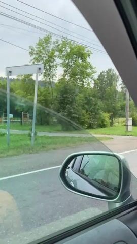 The video shows a view from inside a car looking out the passenger window. The car appears to be in motion, traveling along a paved road. To the left, there is a grassy area with trees and a utility pole. In the distance, a red structure, possibly a playground set, is visible through the trees. The sky is overcast, suggesting cloudy weather. The car's side mirror is visible in the foreground, reflecting the interior of the car and the view behind.