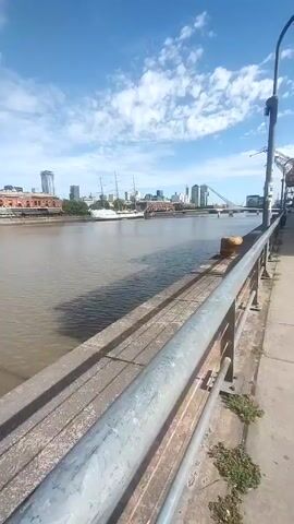 The video shows a waterside promenade on a sunny day. The camera is positioned along a concrete pathway that runs parallel to a river or bay. A metal railing separates the pathway from the water, and a street lamp stands nearby. Across the water, there are buildings, including a tall ship and a bridge.

On the right side of the pathway, there are modern buildings with glass facades. Tables and chairs suggest a restaurant or cafe area. In the distance, two people are jogging along the pathway.

The sky is blue with scattered white clouds, creating a bright and clear atmosphere. There are some industrial cranes in the background, possibly remnants of dock infrastructure.