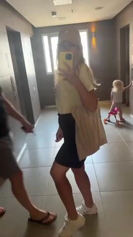 The media file is a selfie video taken in a hotel hallway, likely in Petah Tikva, Israel. The subjects are a couple posing for the camera, with a child playing in the background. The woman, in the foreground, holds up a yellow smartphone to take the selfie. She is wearing a yellow t-shirt, a black skirt, a beige baseball cap, and white sneakers. The man beside her is wearing a black t-shirt, grey shorts, and black flip-flops. He holds up a small, colorful object, possibly a souvenir or toy. In the background, a young child, wearing a pink outfit, is playing with a yellow toy on wheels. The hallway has tiled floors, wood-paneled walls, an elevator door, and light fixtures on the wall. The lighting suggests it is daytime. The overall mood appears casual and playful. There is no visible text in the media file.