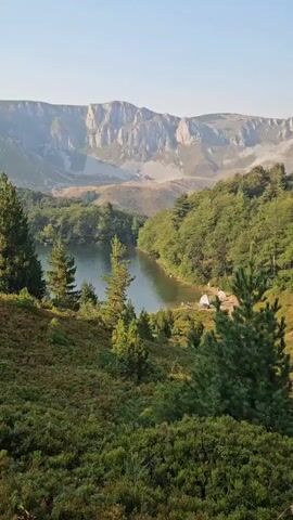 The video shows a wide shot of a mountainous landscape with a lake and a campsite. The mountains in the background are rugged and rocky with some vegetation. The lake is calm and reflects the surrounding trees. The campsite is located on the shore of the lake and consists of a few tents and some people who are not clearly visible. The foreground is filled with lush green trees and bushes. The overall scene is serene and picturesque.