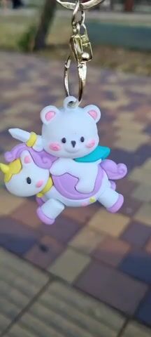A close-up shot shows a keychain dangling with a white teddy bear riding a white unicorn with purple and pink accents. The background is blurred, with a pattern of interlocking paving stones and some greenery. The light is natural, indicating it's outdoors during the day. The keychain is attached to a gold-colored clasp.