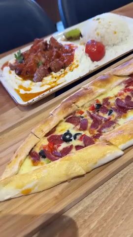 The video pans across a table setting with two plates of food. In the foreground, a long, boat-shaped flatbread topped with melted cheese, pepperoni, black olives, and red bell peppers is presented on a wooden board. In the background, a white plate holds a portion of white rice garnished with small yellow specks, a bright red tomato, and a small piece of green garnish. To the left of the rice, there is another dish with what appears to be stewed meat in a red sauce. The lighting suggests an indoor setting, possibly a restaurant.