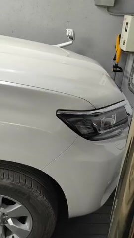 The video shows a close-up, side view of a white SUV. The camera pans from the front fender and headlight area, past the side mirror, to the side of the vehicle's body and window. The vehicle is in what appears to be an indoor garage or workshop, with grey walls and fluorescent lighting visible in the background. There is a partial view of a tire in the foreground.