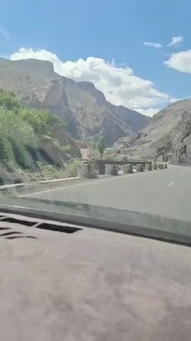 The video, shot from the dashboard of a car, features a scenic drive through mountainous terrain. The road is paved and bordered by concrete barriers, which have painted zebra stripes on them. On the right side of the road, a dirt path ascends the mountain, revealing glimpses of trees, buildings, and a power line. The mountainside is rugged with sparse vegetation and rock formations. The sky above is blue with scattered clouds. The interior of the car is visible, with a textured dashboard covering the lower part of the frame. There is background music playing.