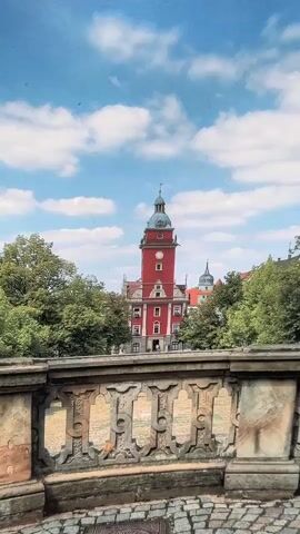 The video captures a scenic view of a red tower with a blue roof, possibly a clock tower, set against a backdrop of lush green trees and a bright blue sky dotted with white clouds. The camera pans slowly from left to right, revealing more of the surrounding architecture, which includes buildings with red-tiled roofs. In the foreground, a stone balustrade with intricate carvings runs along the bottom of the frame, adding a touch of historical charm. The cobblestone ground is visible beneath the balustrade. The overall impression is one of a picturesque European town on a clear, sunny day.