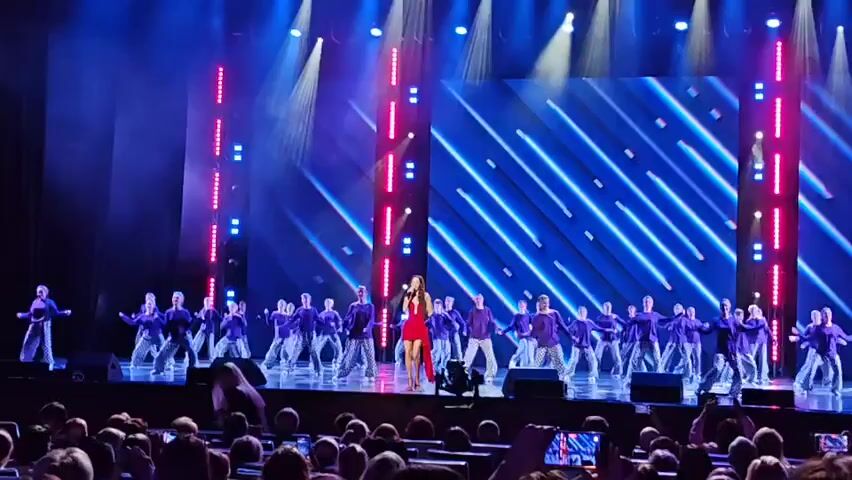 A female singer in a red dress stands center stage, holding a microphone and singing. She is surrounded by a large group of dancers wearing purple tops and patterned pants. The stage is lit with blue and pink lights, and a large screen in the background displays blue diagonal lines. The audience is visible in the foreground, with many people holding up phones to record the performance.