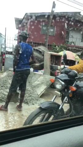 A street scene showing construction materials, including bags of cement and piles of sand and gravel, being worked on by several people. There are temporary market stalls with umbrellas, and buildings in the background. The scene appears to be a construction site or an area where building supplies are sold and processed.