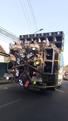 The video shows a large, elaborate sound system mounted on the back of a truck, driving down a street. The sound system is comprised of numerous large speakers stacked vertically and horizontally. In the center of the rear of the truck, a large, multi-spoked wheel is adorned with numerous colorful LED lights, rotating and flashing in sequence. There are also spotlights mounted on the sides of the sound system. The truck is moving forward on a paved road, and the view is from behind and slightly to the side of the truck. The environment appears to be a town or city, with buildings and power lines visible in the background. Sunlight suggests it is daytime.