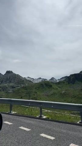 The video is a vertical, medium-long shot of a mountainous landscape. The foreground shows a grey asphalt road with a metal guardrail. To the right of the road, grass and some purple wildflowers grow. In the middle ground, green, rolling hills rise up, dotted with rocks and some trees. Two tall, dark metal electrical towers are visible on the hills. In the background, jagged, rocky mountains with patches of snow are visible against a cloudy, grey sky. The shot appears to be taken from a moving vehicle, possibly a car. There are no people or animals visible in the frame.