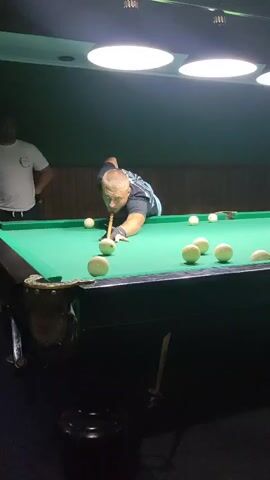 A man is playing pool. He is bent over a green pool table with a cue stick in his hands, about to strike a white ball. There are several other white balls scattered on the table. Another man stands behind him to the left, observing. The room is dimly lit by overhead lights, and the walls are covered in dark green fabric.