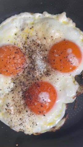 A close-up, overhead shot shows three fried eggs in a black frying pan. The eggs are cooked sunny-side-up, with bright orange yolks. They are seasoned with black pepper and what appears to be salt. The egg whites are mostly white and set, with some slightly browned edges on one side of the eggs. The pan appears to be on a stove as there is a light sizzling sound.