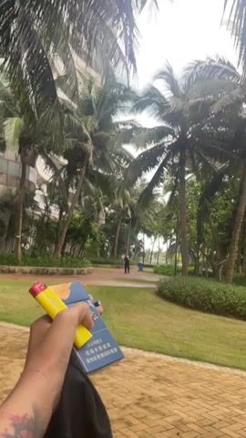 A first-person perspective video pans from a hand holding a yellow vape and a dark blue object with text to a view of a modern building and palm trees. A person with blond hair, facing away from the camera, walks across a grassy area.