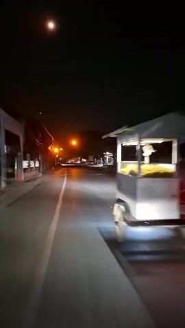 A nighttime street scene with a mobile food cart. The cart, attached to a motorcycle, is illuminated by bright lights, showcasing a large quantity of food, possibly popcorn, in a display case. The motorcycle and cart are moving away from the viewer down a paved road. The road is lined with walls and some sparse vegetation. Streetlights and a bright full moon are visible in the dark sky. The overall atmosphere is that of a late-night street vendor operating in a semi-urban or suburban environment.
