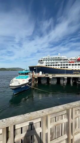 This is a vertical video of a large white and blue cruise ship docked at a pier. The ship has "HAVILA KYSTRUKTEN" written on its side. A smaller teal and white boat is tied to the larger ship. The pier is made of weathered wood planks and has a wooden railing. In the background, there are green, tree-covered hills and a bright blue sky with white clouds. The water is dark and calm. A shadow of a person is visible on the wooden pier in the foreground.