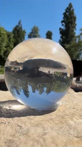This is a close-up, low-angle shot of a clear glass sphere resting on a rough, beige-colored stone surface. The sphere is partially filled with water, creating a lens that refracts the light and inverts the scene behind it. The top half of the sphere is dry and captures the upper portion of the landscape, including the bright blue sky with a few wispy white clouds and the tops of tall green trees. There's a strong glare reflecting off the top surface of the sphere, indicating a sunny day.

The bottom half of the sphere, filled with water, acts as a lens, showing an inverted reflection of the scene below. This includes the green trees and the blue sky, creating a striking visual contrast. A dark, amorphous object is submerged in the water, its shape indistinct. The overall setting appears to be outdoors, likely a park or garden area given the presence of trees and the natural stone. The lighting suggests it is daytime, with the sun high in the sky. The scene is serene and visually captivating due to the distortion and reflection within the glass sphere. There is no visible text in the media file. The context provided places this scene in Beverly Hills, United States of America.