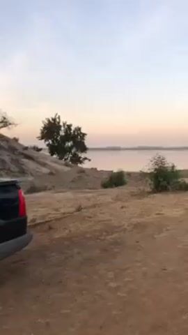 The video depicts a tranquil lakeside view at dusk. A dark-colored vehicle is parked in the foreground on a sandy, dirt-covered area with sparse vegetation. The focus shifts to a calm lake extending into the distance, where a small boat with two people is visible. In the background, there are distant hills or landmasses. The sky displays a gradient of soft colors, indicative of a sunset or sunrise.