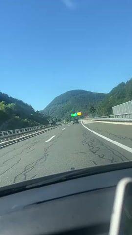 The video shows a view from inside a car driving on a highway. The road is a multi-lane highway with a clear blue sky above. On either side of the highway, there are green, tree-covered hills. In the distance, there are two overhead signs with traffic lights. The sign on the left has a green light, and the sign on the right has a red light. A dark-colored car is visible in the distance, driving in the same direction as the camera car. The dashboard of the car is visible in the foreground, and a portion of a smartphone can be seen in the lower right corner of the frame. The highway appears to be in good condition, with white lines marking the lanes. There are some cracks visible on the road surface. There are also several green signs with white text and symbols along the side of the road. One sign on the left shows a white pedestrian icon and the number 800. Another sign on the right shows a speed limit symbol with the number 100.