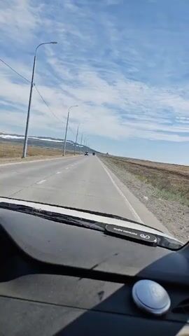 The video shows a first-person view from a vehicle driving on a highway. The road is asphalt with white lines marking the lanes. On the right side of the road, there is a dry, grassy embankment and sparse vegetation. In the distance, mountains with snow-capped peaks are visible. The sky is blue with scattered white clouds. Several streetlights are visible along the side of the road, and other vehicles can be seen in the distance. The dashboard of the vehicle is visible in the foreground, with a phone mount and a circular object. The scene appears to be a rural or remote area.