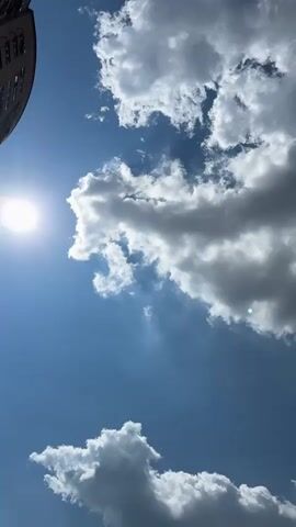 The video shows a time-lapse of clouds moving across a blue sky. The sun is visible, shining brightly and casting shadows. A portion of a building is visible on the left side of the frame. The clouds are white and puffy, and they appear to be moving from left to right across the sky. The video is shot from a low angle, looking up at the sky.
