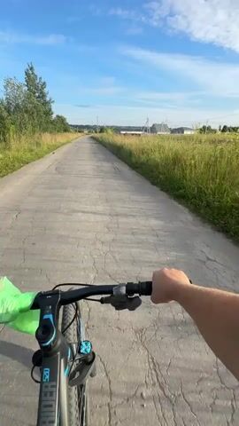 A first-person perspective video shows a person riding a bicycle down a cracked asphalt road. The road is bordered by tall green grass and trees on the left, and a field of grass on the right. In the distance, there are houses and power lines under a bright blue sky with scattered white clouds. The sun is visible, shining through the clouds. The rider's hands are visible on the handlebars, and a green bag is attached to the handlebars. The camera angle is tilted slightly upwards, capturing the sky and the surroundings.
