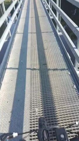 A first-person perspective video shows a person walking across a metal pedestrian bridge. The bridge has metal railings on either side and a grated metal walkway. The video pans slightly to the left, revealing a body of water below with trees and vegetation on the far bank. The sun casts shadows across the bridge, indicating it is daytime. The person's footwear, a dark boot, is visible at the bottom of the frame. The person is walking at a steady pace towards the other end of the bridge. The overall scene is one of outdoor activity on a pleasant day.