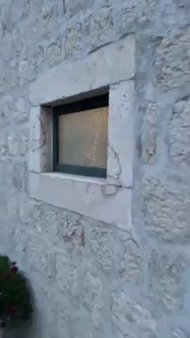The video shows a close-up of a small, rectangular window in a stone wall. The window has a green frame and textured glass that obscures the view inside. Silhouettes of two people are visible inside the room, possibly moving around. The camera zooms in slightly on the window before pulling back to show more of the stone wall and a doorway next to the window. The video appears to be taken outside in daylight.