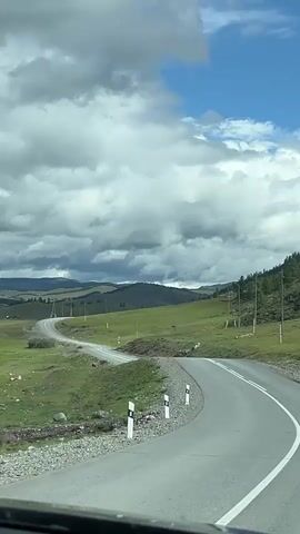 The video shows a curved asphalt road in a mountainous, rural landscape. The road has a double white line down the center and is bordered by gravel on the left and green grass on the right. Several utility poles are visible along the right side of the road. In the distance, rolling green hills with some scattered trees are visible under a cloudy sky with patches of blue. The camera appears to be mounted inside a vehicle, looking forward. There are no people or other vehicles visible in the frame. The overall impression is a scenic drive through a natural environment.