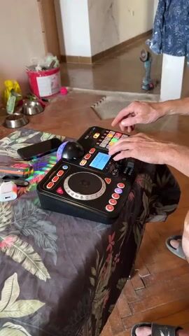 A person's hands are visible operating a black DJ controller with a circular platter and various buttons and knobs. The controller is placed on a surface covered with a patterned cloth featuring large green leaves and red flowers. To the left of the controller, a smartphone is visible, along with some colorful items, possibly toys. In the background, a person's legs and feet are visible, wearing sandals. The environment appears to be indoors, with wooden flooring and a glimpse of what looks like a room with some construction or renovation materials, including a power tool and paint cans, in the distance. The lighting suggests an indoor setting.