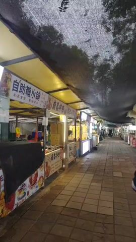 The video depicts a lively outdoor food street or night market, likely in Shapingba District, China. The scene is shot from a first-person perspective, moving forward along a paved walkway made of light-colored bricks.

On both sides of the walkway, numerous food stalls are set up, each featuring bright yellow awnings and illuminated by LED strip lights. A dark, mesh-like material covers the entire walkway overhead, suggesting an evening or night setting.

The stalls display various food items and menus, predominantly in Chinese text. Prominently visible signs include "糖水铺" (Tángshuǐ pù), indicating a dessert or sweet soup shop, and multiple signs for "南昌拌粉" (Nánchāng bànfěn), which translates to Nanchang Mixed Rice Noodles, and "锅巴拌饭" (Guōbā bànfàn), or Crispy Rice Mixed with Rice. Other signs show prices like "10元" and "15元" (10 and 15 yuan).

A few people can be seen walking in the distance further down the market path, and the feet of the person recording are visible in the foreground. The stalls appear to be equipped for cooking and serving food, with some showing stacks of boxes. The overall atmosphere is bustling and inviting, typical of an Asian night market.