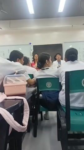 The video shows a classroom setting where a group of students, all wearing white tops, are seated at desks facing a front area. Two individuals, a woman in a red top and a man in a white lab coat, appear to be lecturing or presenting. One student in the foreground, partially obscured, is wearing glasses and has their hair in a bun. Other students are also facing the front, some with their heads down. A pink storage container and a pink bag are visible on a desk in the foreground. The room is brightly lit by fluorescent ceiling lights. The whiteboard in the background has some writing on it, and a drip stand is visible. The overall impression is of a medical or science-related educational environment.