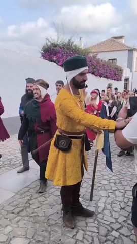 A group of people in medieval-style clothing are walking down a cobblestone street. In the foreground, a man in a yellow tunic with intricate patterns, dark leggings, and brown boots is holding a stick and a blue and yellow flag. He has a beard and is wearing a green and white hat. He is interacting with someone off-camera. To his left, another man in a red and green tunic with brown leggings and boots is carrying a lute. Behind them, several other individuals are dressed in historical attire, including a man in a white robe with a red cross, and women in long dresses. A white building with purple flowers growing on it is visible in the background. The sky is cloudy.
