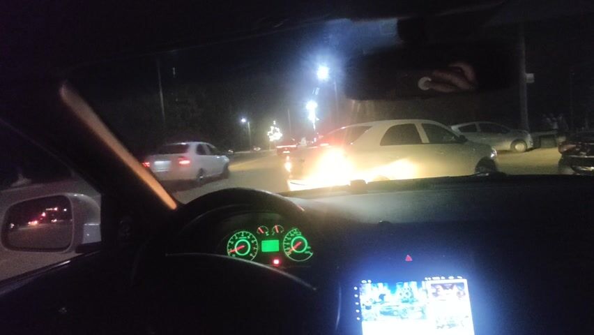 The video shows a view from inside a car driving at night. The car is on a road with other vehicles. There are streetlights illuminating the road. The dashboard of the car is visible with a digital display. The side mirror shows headlights of vehicles behind the car. Trees line the side of the road.