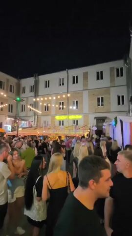The video shows a crowded outdoor scene at night, likely a market or festival. A multi-story building with light-colored walls and windows forms the backdrop. String lights are hung across the space, and a neon sign with yellow lettering is visible on the building. People are densely packed, milling about and interacting. Some individuals are in focus, while others are blurred due to movement. Various stalls or vendors are suggested by canopies and tents. The overall atmosphere appears lively and social.
