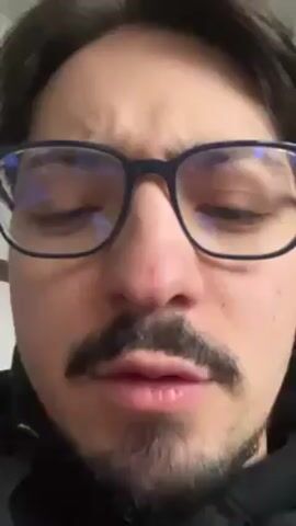 The video shows a man's face in close-up. He has dark hair, a mustache, and is wearing glasses with dark frames. His facial expression is neutral, and he appears to be looking slightly downward. The background is a plain, light color, and the lighting is even. The man's face is the primary focus of the video.