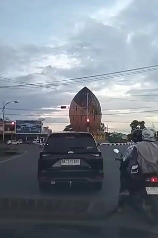 This video captures a street scene in Sigli, Indonesia, likely at dusk given the muted lighting and cloudy sky. The primary focus is a distinctive, large, acorn-shaped sculpture situated at an intersection with traffic lights. A black SUV is stopped at the red light, with its license plate visible as "BK 1189 AFF." To the right of the SUV, a motorcyclist wearing a helmet and a plaid shirt is also stopped at the light, his backpack clearly visible. Another motorcyclist is visible further down the road to the left, also stopped at a red light.

In the background, a building with a large advertisement banner is visible, and beyond that, a few houses with colorful roofs dot the landscape. Overhead, power lines crisscross the sky, a common sight in urban and semi-urban environments. The sculpture, with its wooden lattice-like design, appears to be a significant landmark. The overall atmosphere is that of a typical traffic intersection during a period of lower light. There is no overt emotional display from the subjects, and the activity is simply traffic waiting at a red signal. The time of day is suggested to be late afternoon or early evening. The word "SIGLI" is visible on a sign near the sculpture.