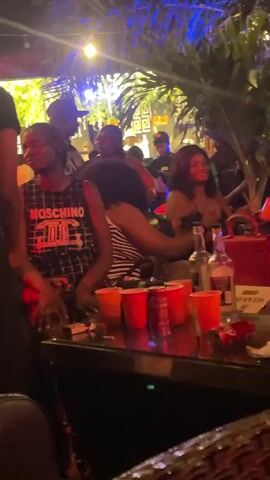 A group of people are gathered around a table at what appears to be an outdoor venue. Red plastic cups and bottles of alcohol are on the table, suggesting a party or social gathering. The lighting is dim, with some blue and green lights creating a festive atmosphere. People are interacting, with some standing and talking while others are seated. The overall scene depicts a lively social event.