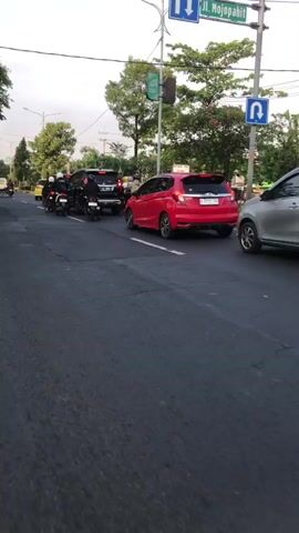 The media file depicts a street scene in Larangan, East Java, likely during the late afternoon or early evening, given the soft, diffused lighting. The sky appears overcast or hazy. The primary focus is a traffic situation on a paved road with visible lane markings.

Several vehicles are present, including a black SUV, a red hatchback, and a yellow vehicle in the distance, possibly another car. A significant number of motorcycles are also on the road, some of which appear to be stopped or moving slowly, suggesting a traffic jam or congestion. Multiple individuals are seen on or around the motorcycles, some appearing to be riders. The presence of a traffic light above the intersection indicates this is likely a junction. Trees line the sides of the road, adding to the urban or semi-urban setting.

The activity is centered around traffic, with vehicles and riders gathered at what seems to be an intersection. The mood appears to be one of typical daily traffic, with no overt signs of distress or celebration, though the density of vehicles suggests a degree of inconvenience.

There is a visible green traffic light signal, and what looks like a sign on a pole with a green emblem, potentially indicating a direction or information. No other discernible text is present in the visible frame. The scene is dynamic, with vehicles and people in motion or paused, creating a snapshot of everyday life in this location.