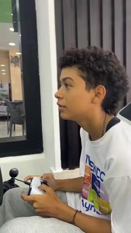 A young person is seated in front of a large screen, holding a game controller and smiling slightly. The screen is displaying a menu with various icons, suggesting a video game interface. The person is wearing a white t-shirt with graphics, grey sweatpants, and a beaded necklace. They have curly hair with a faded haircut. The screen and game controls are mounted on a black metal frame. The environment appears to be inside a brightly lit commercial space, possibly a store or arcade, as seen through the glass windows behind the setup, with tables, chairs, and signage visible in the background. Artificial grass is covering the platform around the base of the seat.