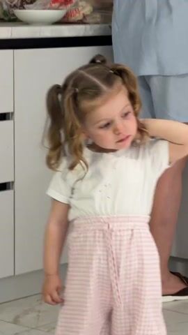A little girl stands in front of a white kitchen cabinet with black drawers. She is wearing a white t-shirt and pink and white checkered pants with a drawstring. Her blonde hair is styled in two pigtails. She rests her right hand behind her head and looks to the right. Behind her, an adult in a blue shirt and gray shorts stands wearing sandals. A white bowl is on the counter, along with other food items.