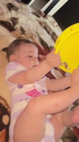 A baby lies on a patterned blanket and plays with a yellow tambourine. The baby is wearing a white and purple striped onesie with a graphic of owls on it. The baby holds the tambourine in its hands and shakes it, producing a jingling sound. The baby's facial expression suggests curiosity and engagement with the toy. The lighting is dim, creating a cozy atmosphere.